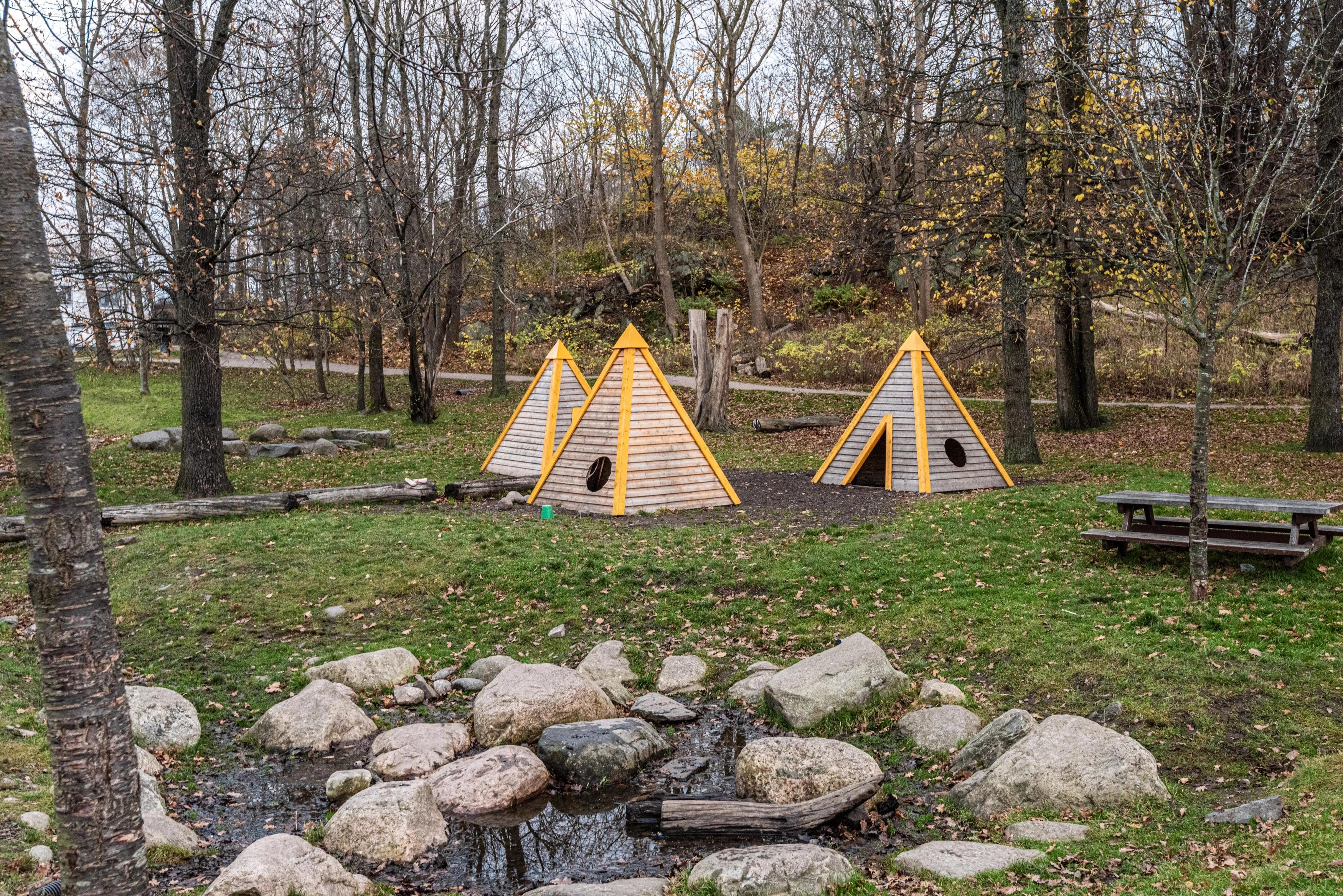 Tři dřevěné pyramidové domečky obklopené stromy, s piknikovým stolem a potůčkem v popředí