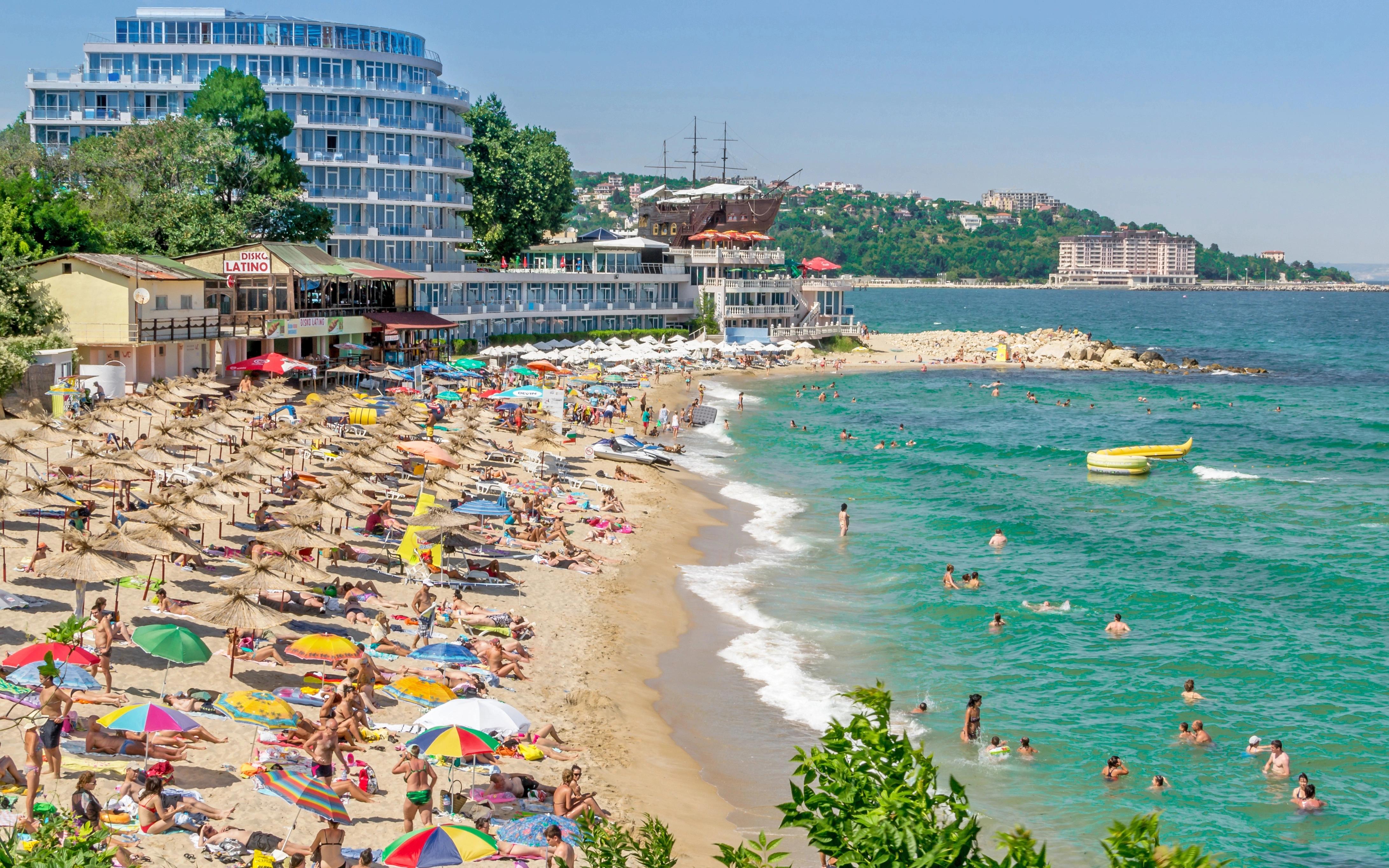 Slunná pláž s lidmi pod slunečníky, tyrkysové moře a hotely v pozadí, barevné deštníky a slaměné stínidla