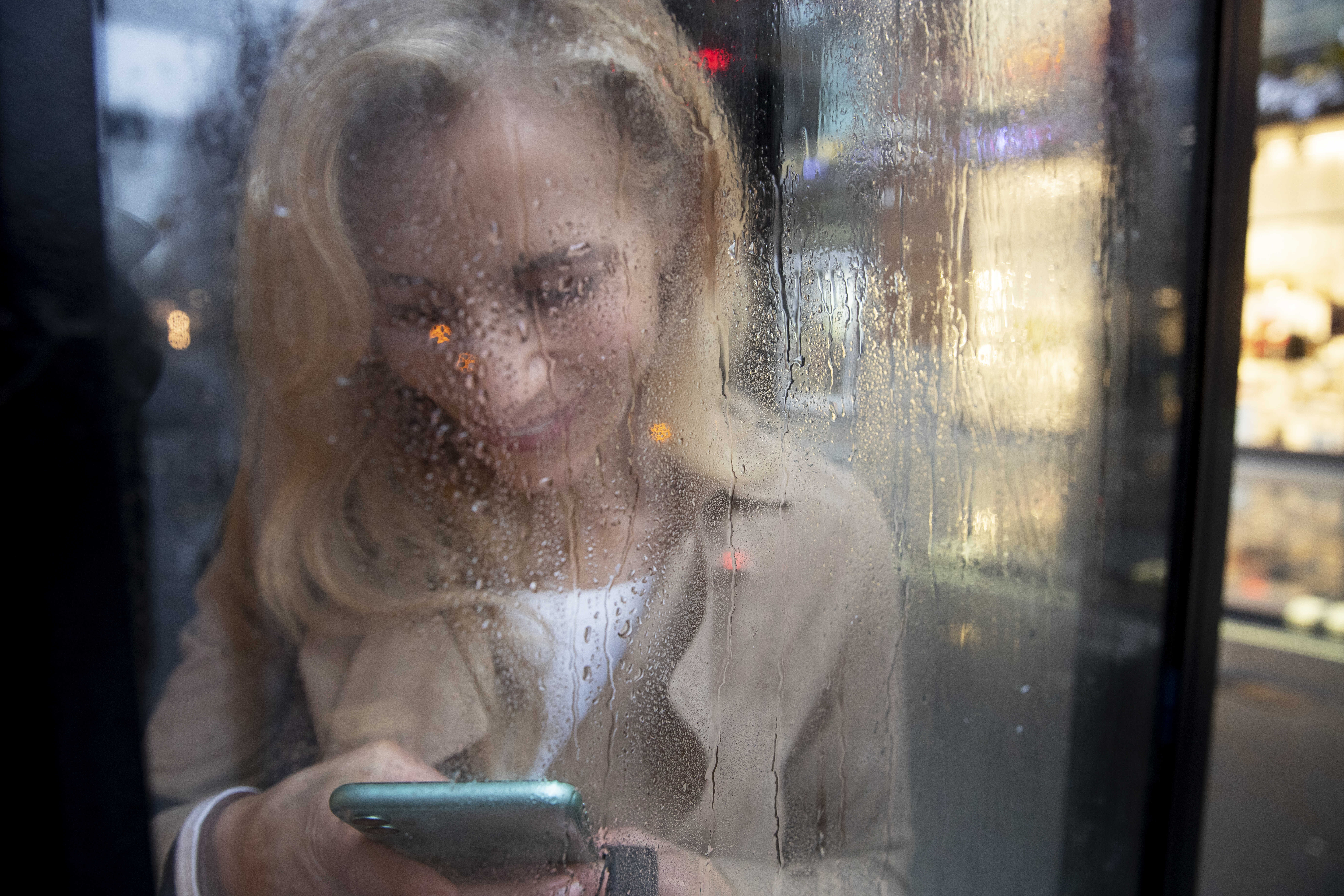 Žena stojící za oknem během deštivého dne, držící mobilní telefon a psaní zpráv