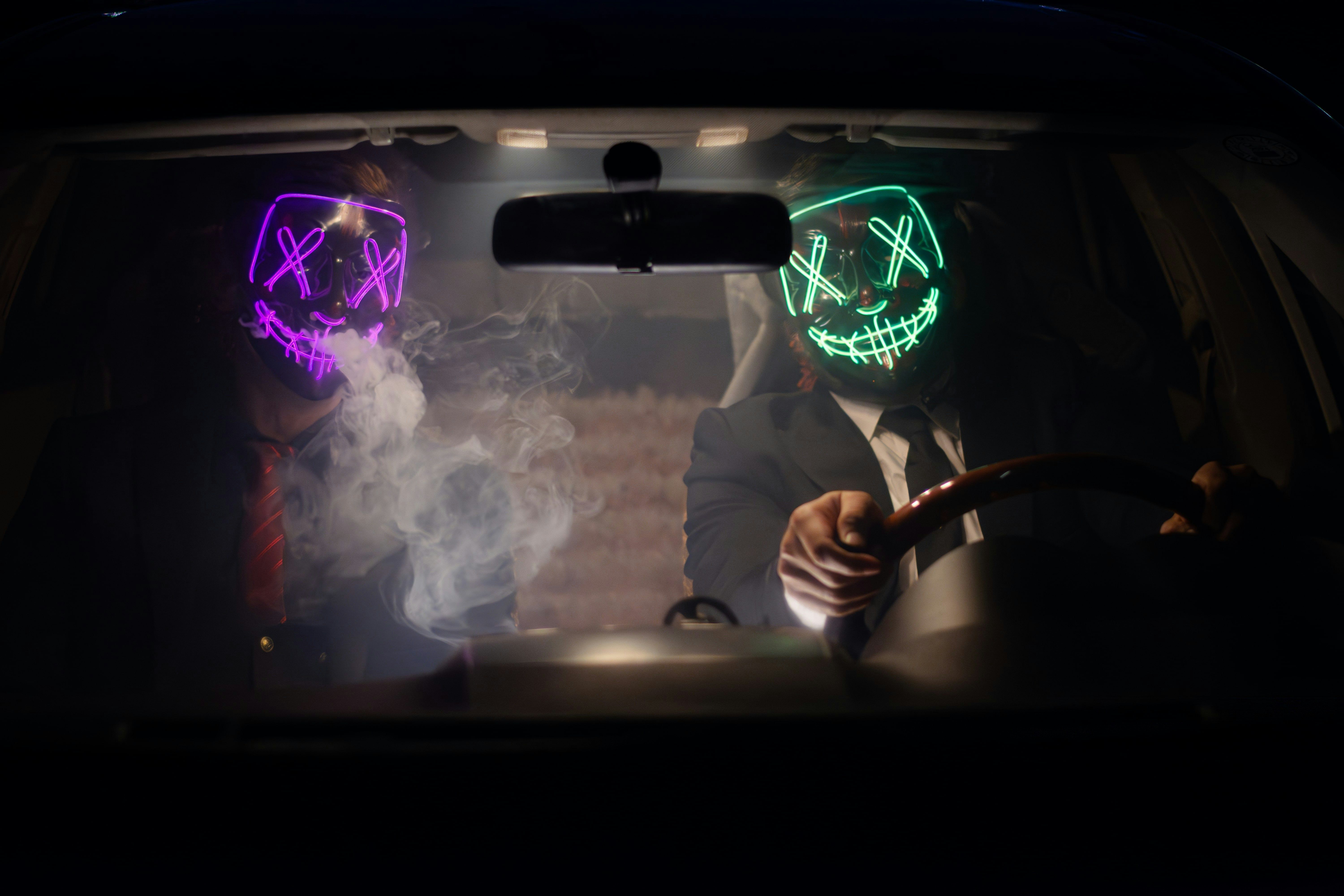 Dva muži sedící v autě s neonovými maskami na obličeji, vytvářející tajemnou atmosféru.