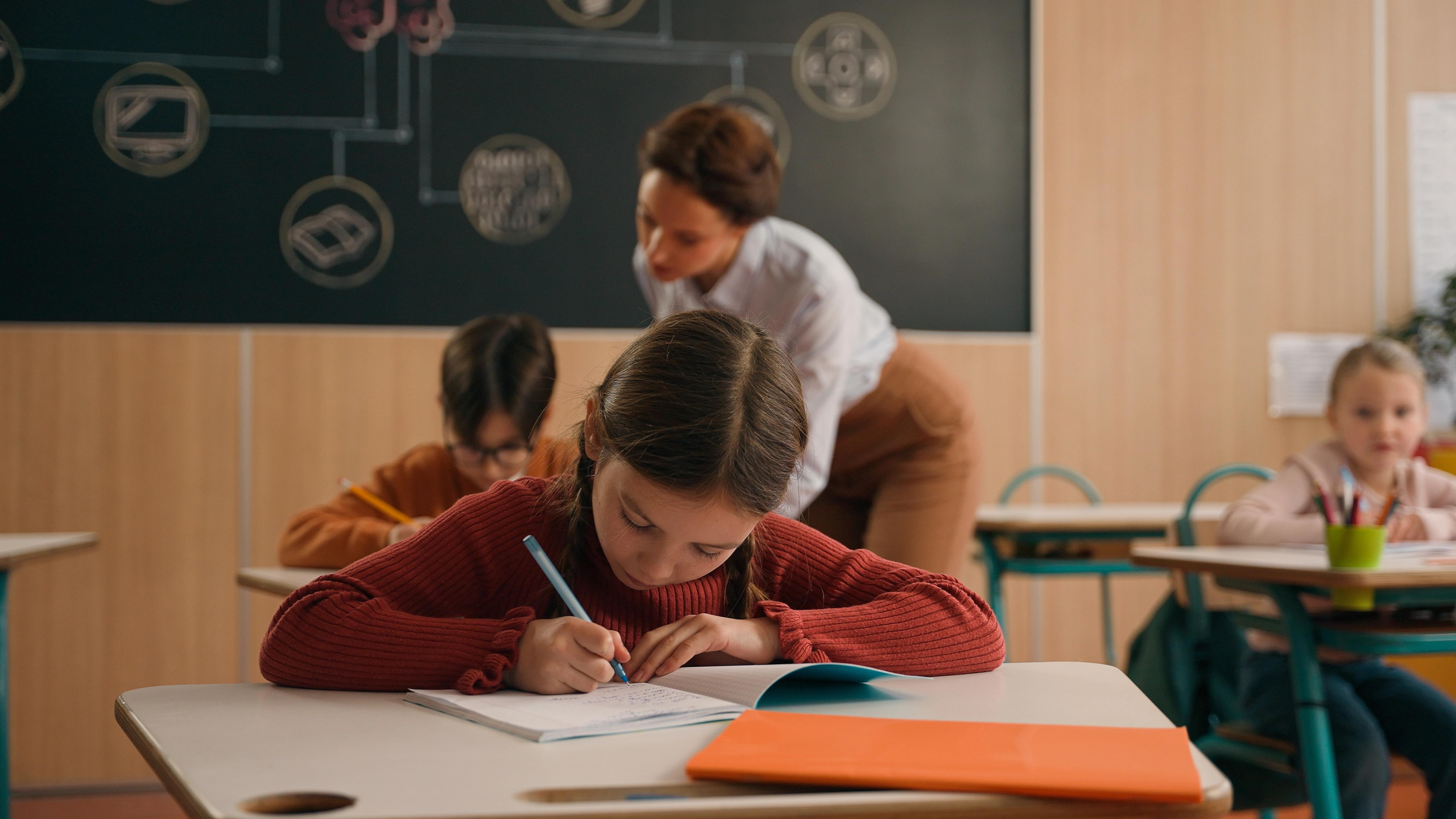 Učitel se sklání k lavici a pomáhá dvěma žákyním při školní práci