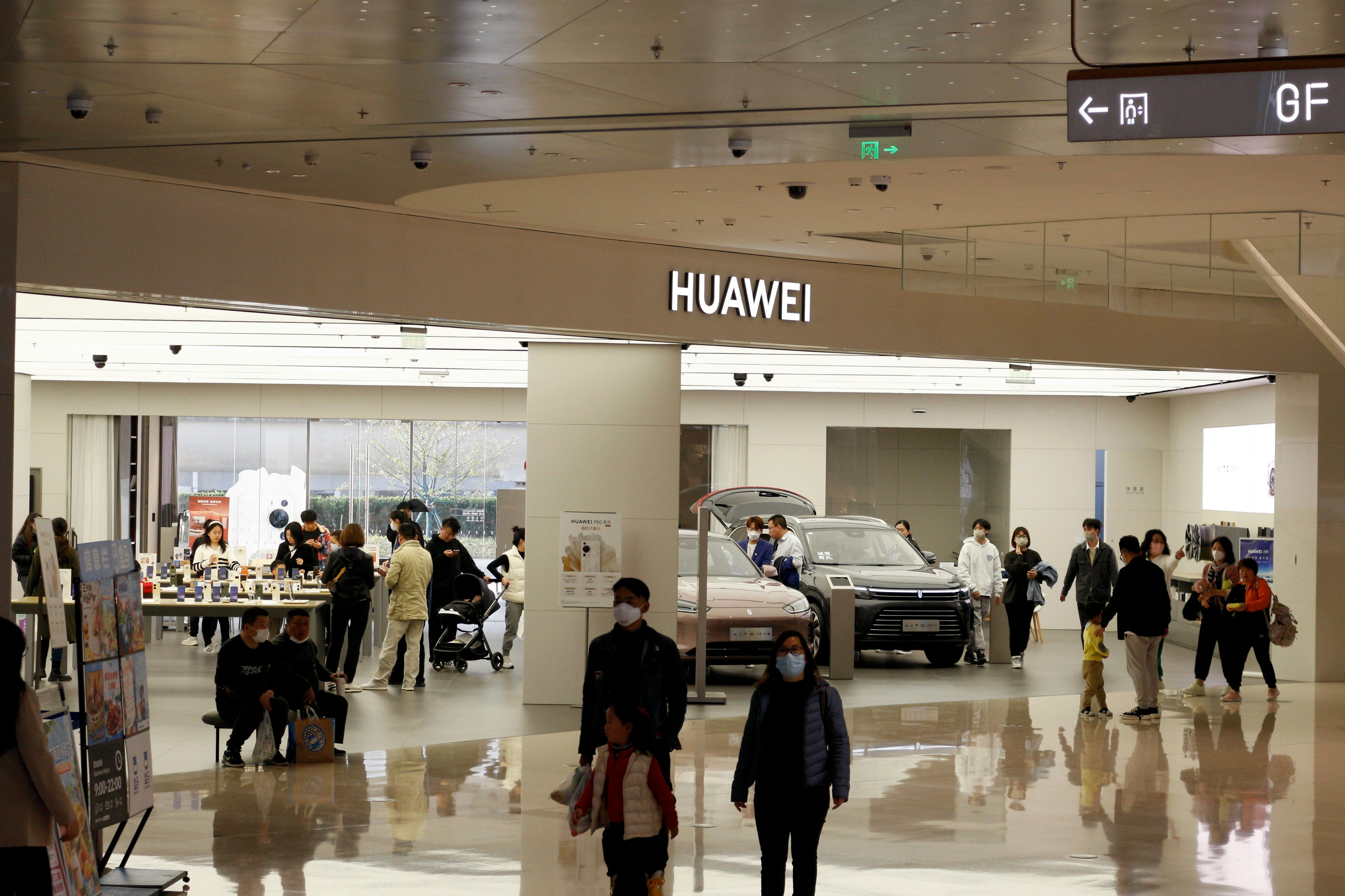 Obchod Huawei v nákupním centru s návštěvníky procházejícími kolem, několik lidí se zastavilo, aby si prohlédli vystavené produkty.