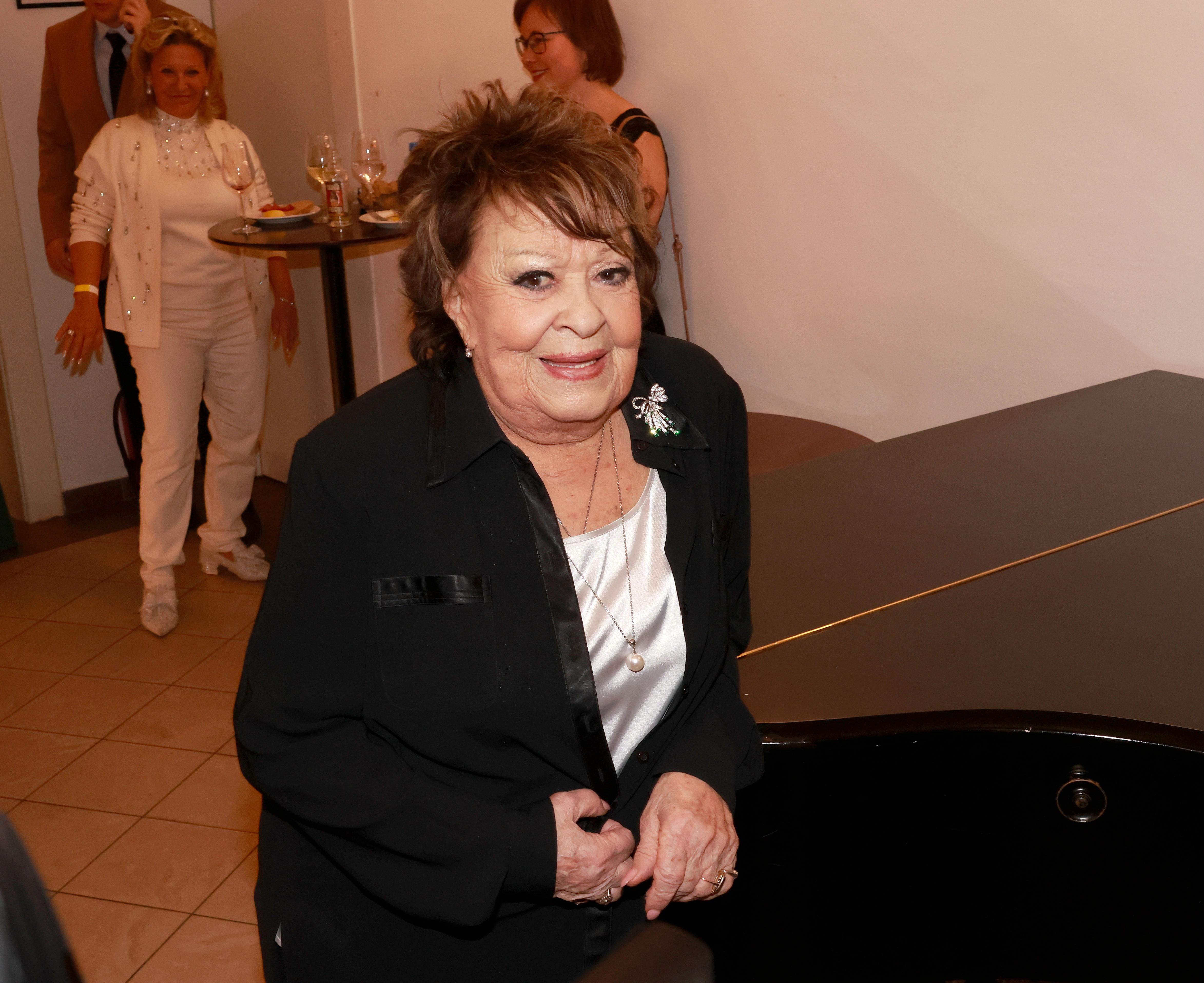 Jiřina Bohdalová v černém sako u klavíru, usmívá se do kamery