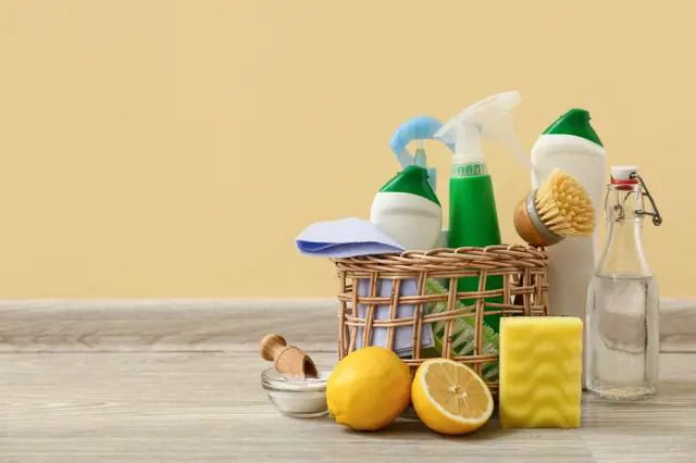 Košík s přírodními čisticími prostředky, citrony, houbičkou a lahvemi na dřevěném stole, béžové pozadí