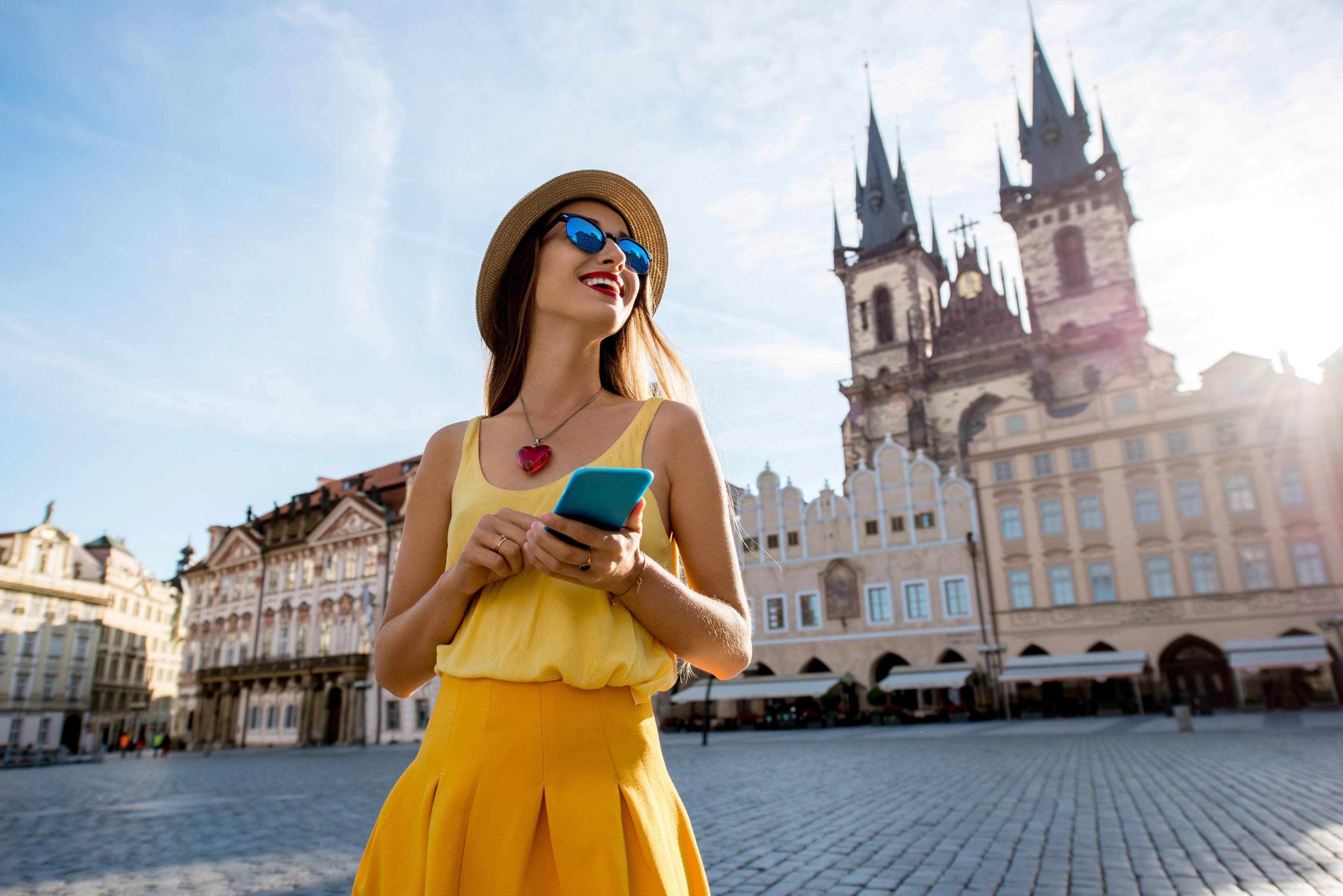 Mladá žena v žlutých šatech stojí na Staroměstském náměstí a drží mobilní telefon